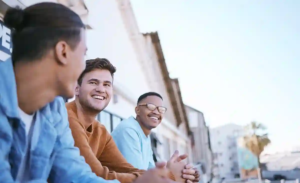 Three casually dressed people sit outdoors near palm trees and buildings, enjoying a relaxed moment in an urban setting
