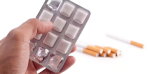 Close-up of a hand holding nicotine gum blister pack, positioned beside scattered cigarettes on a white surface—visually contrasting smoking cessation support with the harmful habit of smoking.