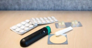 Assortment of medical items on a wooden surface including tablets, capsules, a black inhaler-like device, a white cylinder, and a round adhesive patch—suggesting healthcare treatment or quitting aids.
