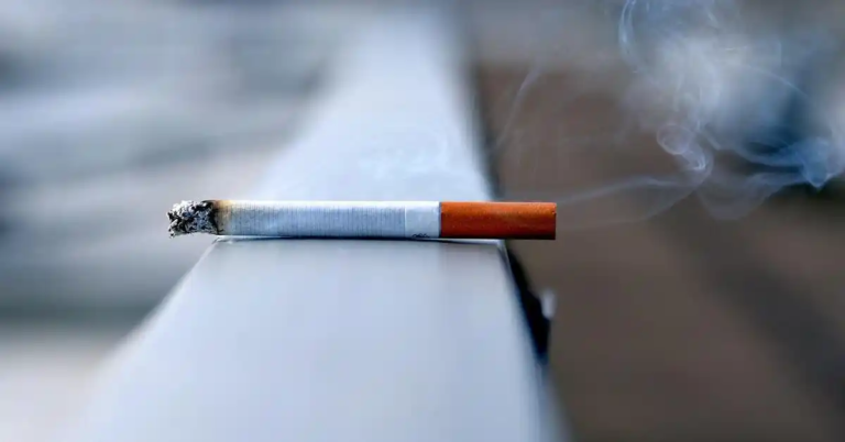 A lit cigarette resting on a metallic surface with smoke rising from its tip, swirling against a blurred background—evoking the fleeting yet persistent nature of the smoking habit.