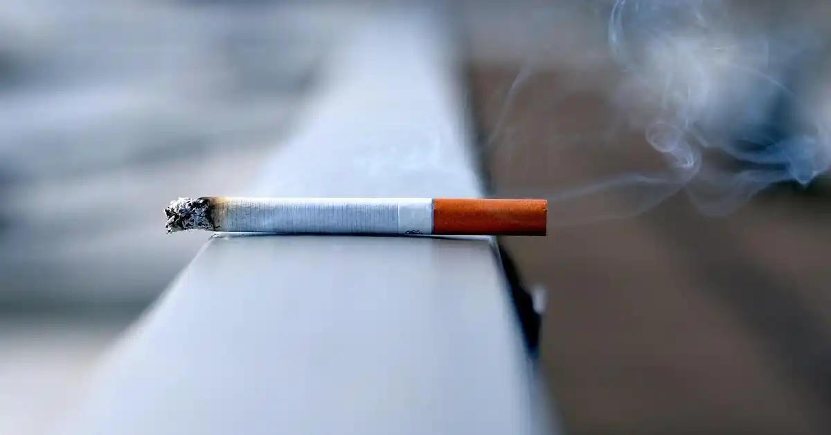 A lit cigarette resting on a metallic surface with smoke rising from its tip, swirling against a blurred background—evoking the fleeting yet persistent nature of the smoking habit.