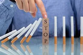 Hand in blue shirt presses a wooden block labeled 'STOP', halting falling cigarettes lined up like dominoes