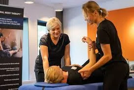 Two practitioners assist a patient lying on a blue therapy table, adjusting the shoulder and arm in a clinical setting with a 'RESET THERAPY' poster in the background