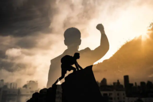 Double exposure image showing a raised fist superimposed over a climber ascending a rocky peak, with a cityscape and bright sun in the background
