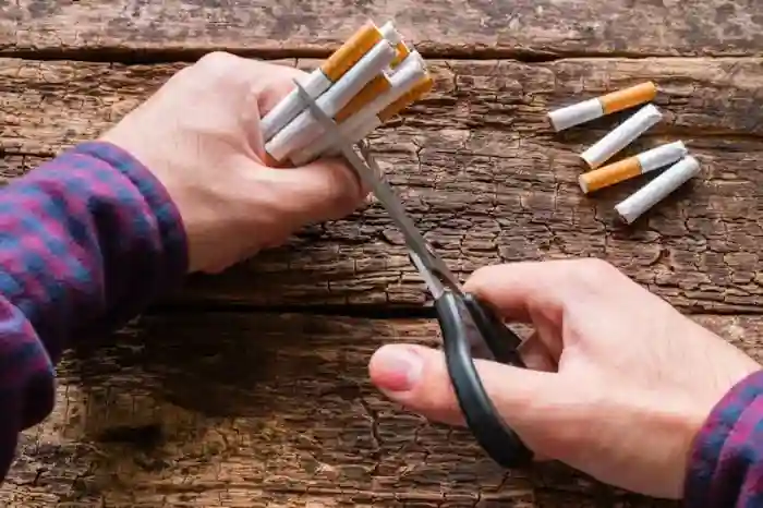 A close-up of hands wearing a plaid shirt using scissors to cut multiple cigarettes over a wooden surface, with cigarette pieces scattered — symbolizing the decisive action of quitting smoking.