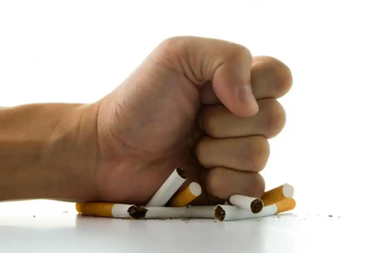 A strong hand clenched into a fist forcefully crushing several broken cigarettes on a white background, with tobacco spilling out — representing the determined act of quitting smoking and rejecting addiction.
